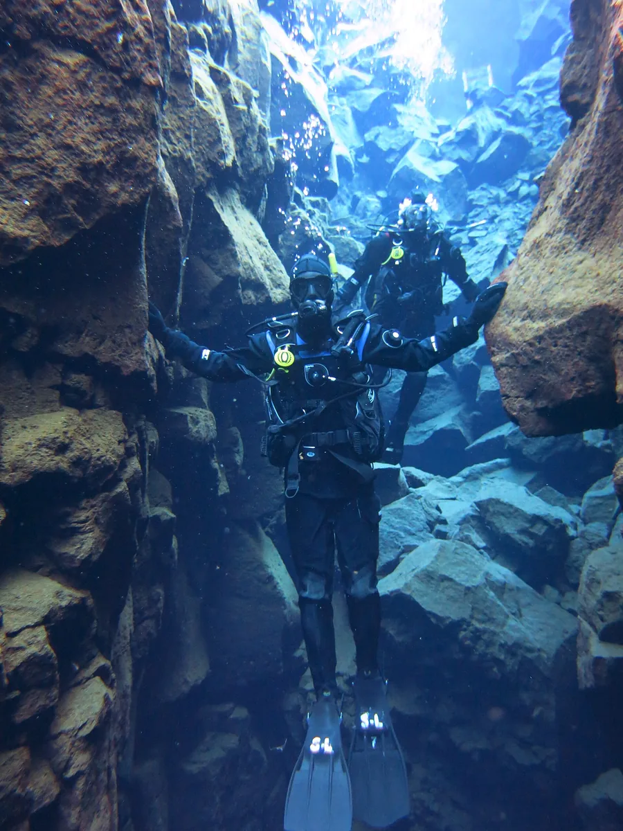 A diver in a dry suit hovers in Silfra between two rock walls in blue water
