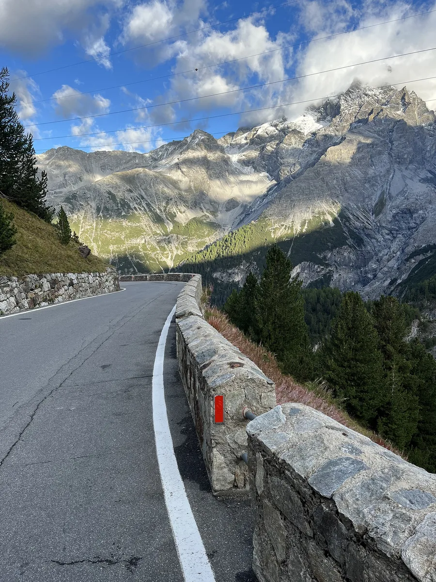A hairpin bend on the Stelvio Pass road in Stelvio National Park with a stone wall, forest and glaciated peaks behind