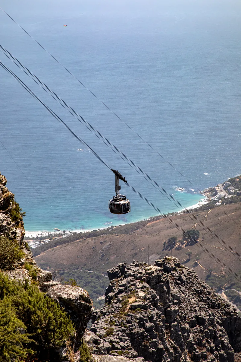 A round car of the Table Mountain cableway hangs from two cables above a rock edge, far below the coast and sea at Camps Bay