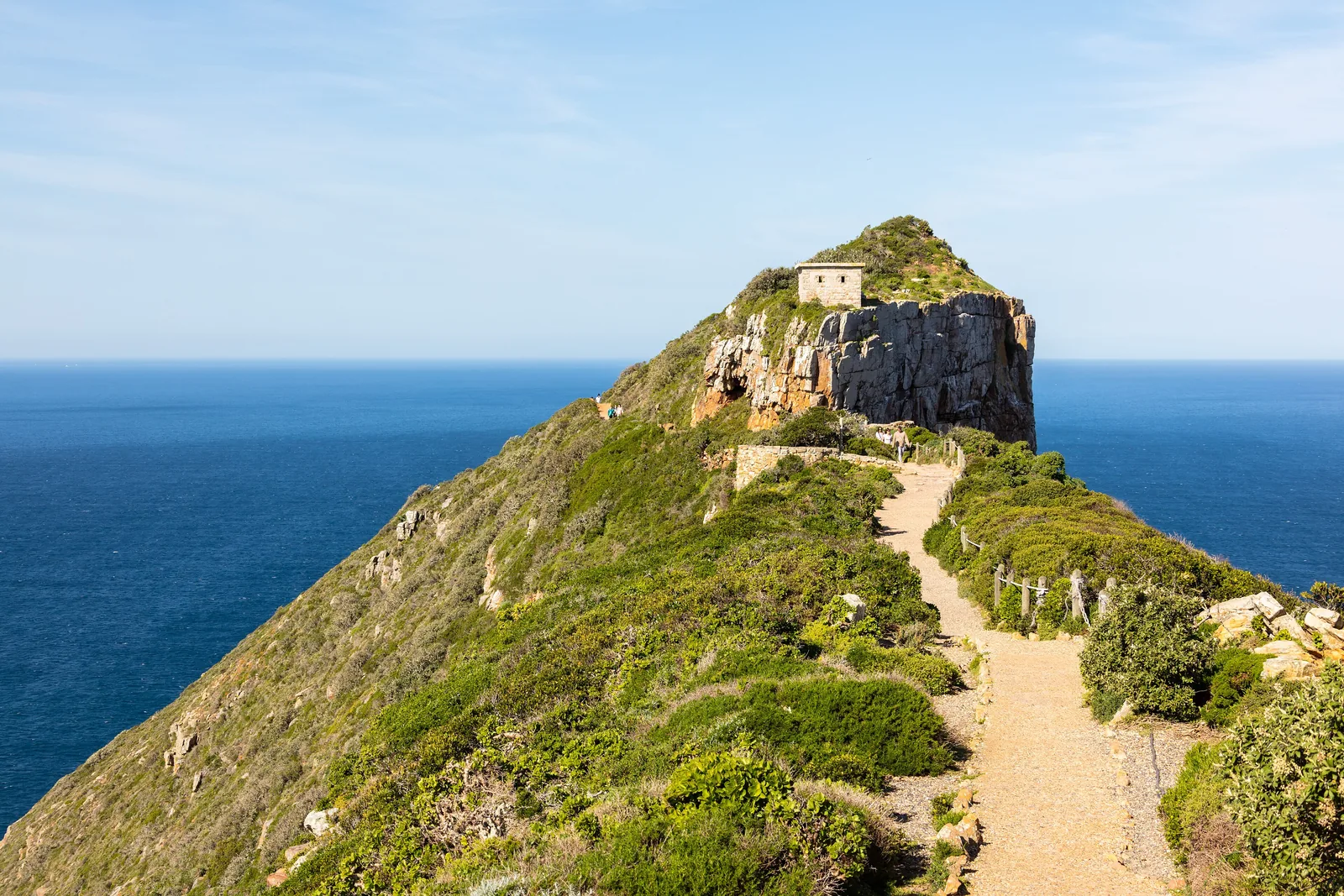 A path runs along a green ridge in Table Mountain National Park to the old lighthouse on the summit of Cape Point, with the sea behind
