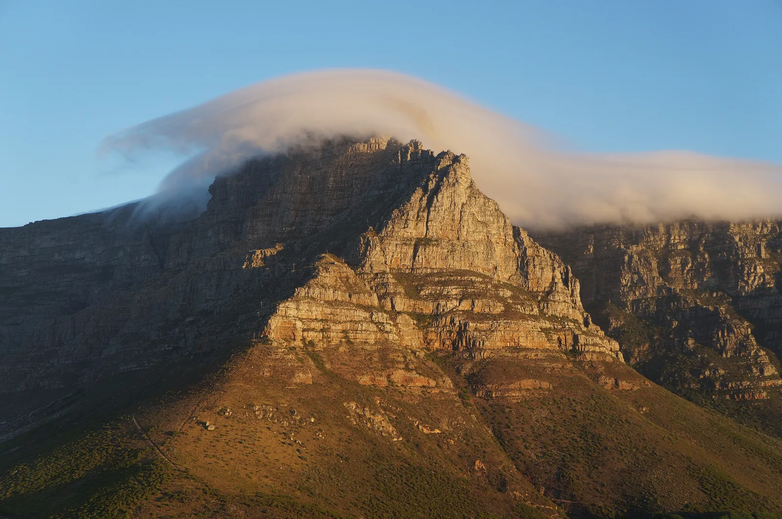 A roll of cloud drifts over the rock faces of the Table Mountain range in evening light, the slopes glowing orange