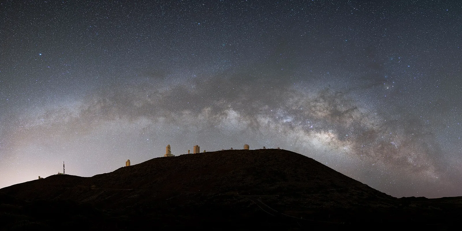 The arc of the Milky Way in Teide National Park over the dark hill of Izaña with the telescope domes