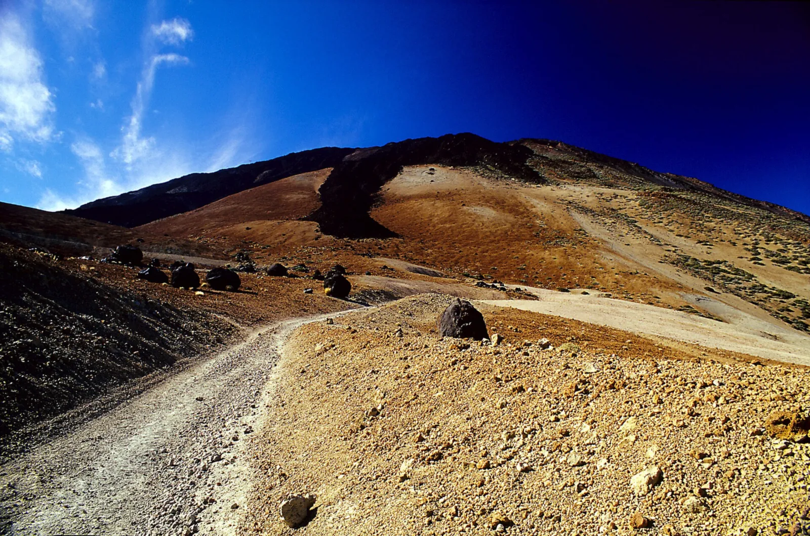A gravel track in Teide National Park across the pale Montaña Blanca, lined with dark, round lava bombs, with a black lava flow behind under a deep blue sky
