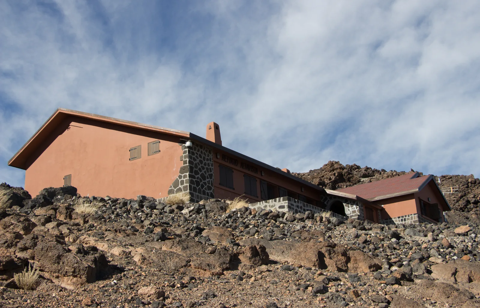 The red Refugio de Altavista in Teide National Park on dark lava ground under wisps of cloud, seen from the ascent trail over Montaña Blanca
