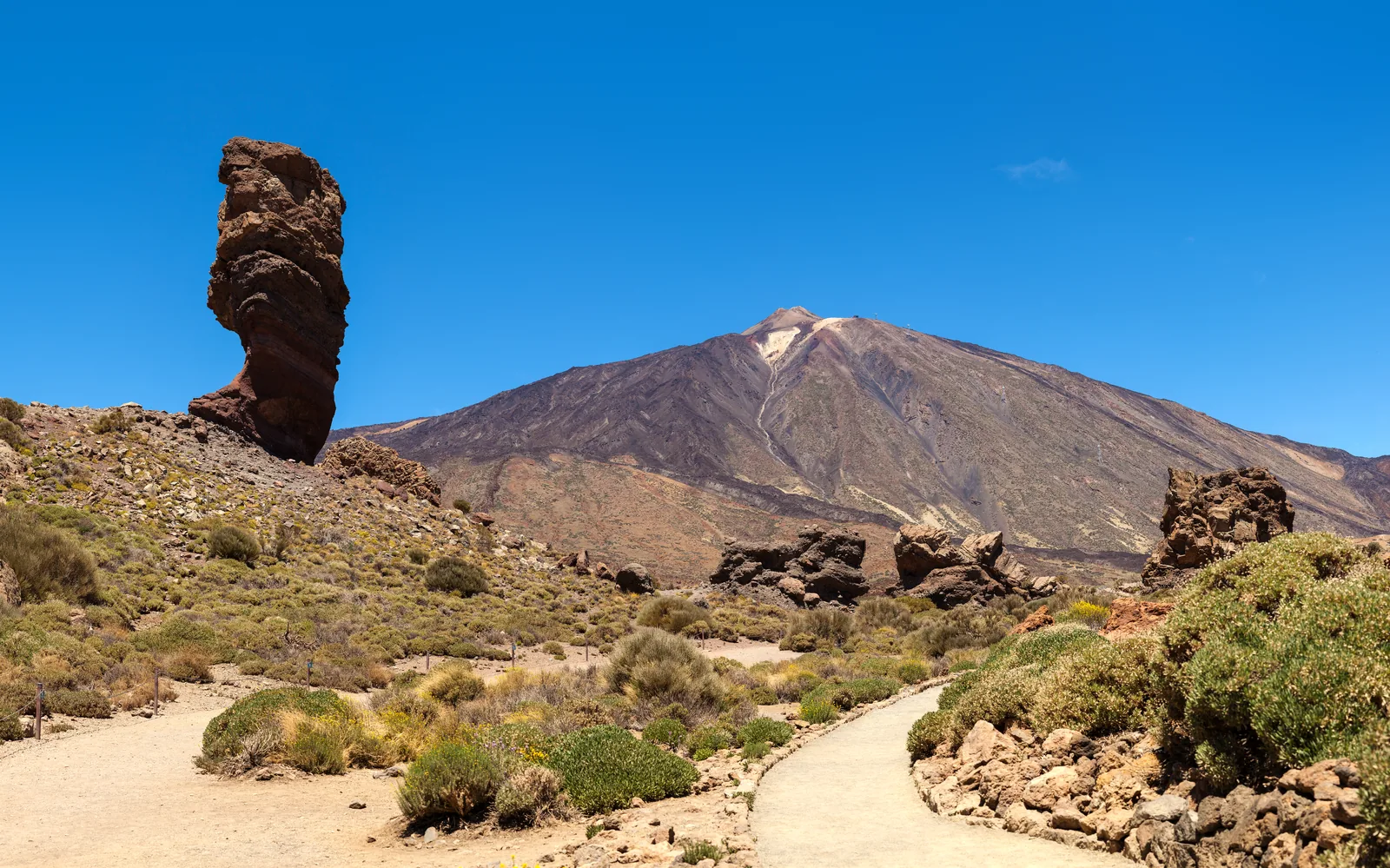 Roque Cinchado, a narrow rock pillar, in front of the cone of Mount Teide in Teide National Park, with the pale path through Las Cañadas in the foreground