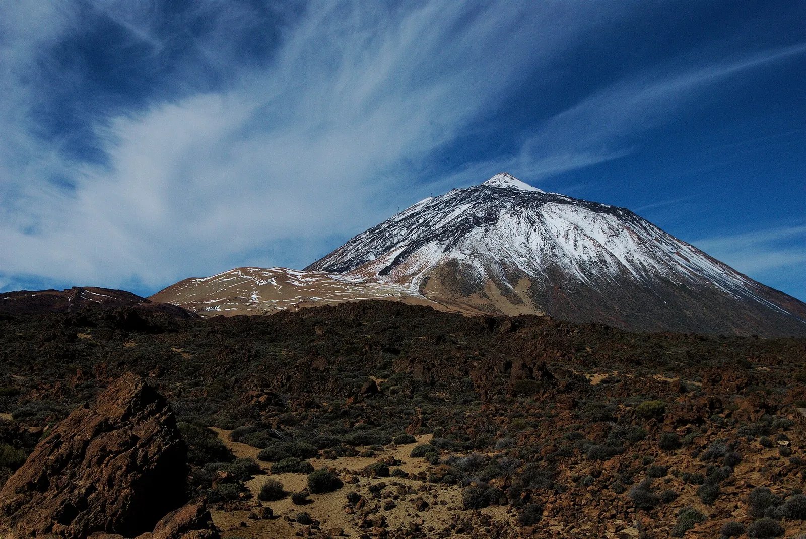 The snow-covered summit cone of Teide and the pale Montaña Blanca in Teide National Park above dark lava fields, with wispy clouds in the sky