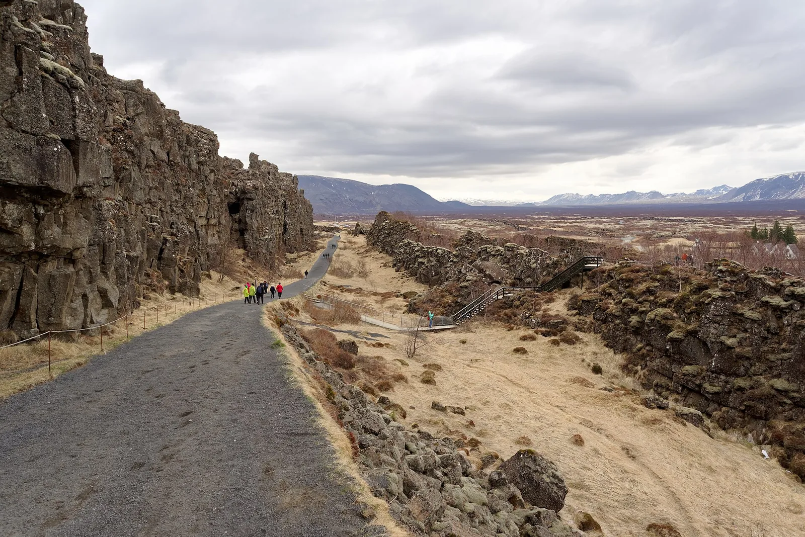 Path through Almannagjá in Þingvellir National Park: the high basalt wall on the left, the rift with dry grass on the right, visitors in the distance
