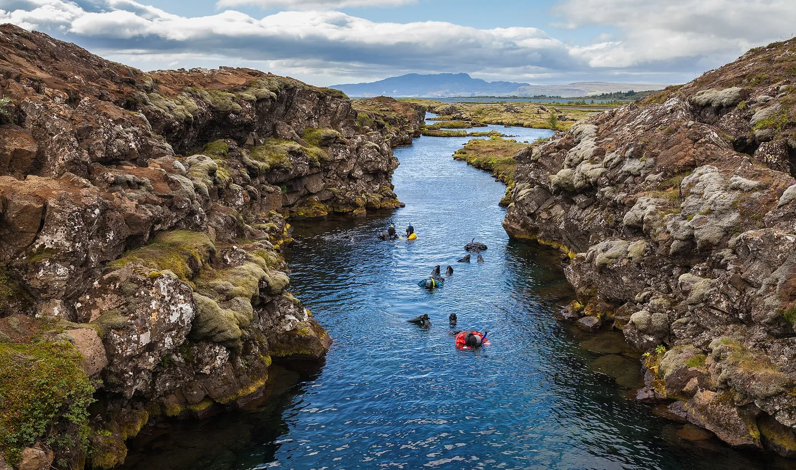 Snorkellers in dry suits drift through the Silfra fissure in Þingvellir National Park between moss-covered lava walls