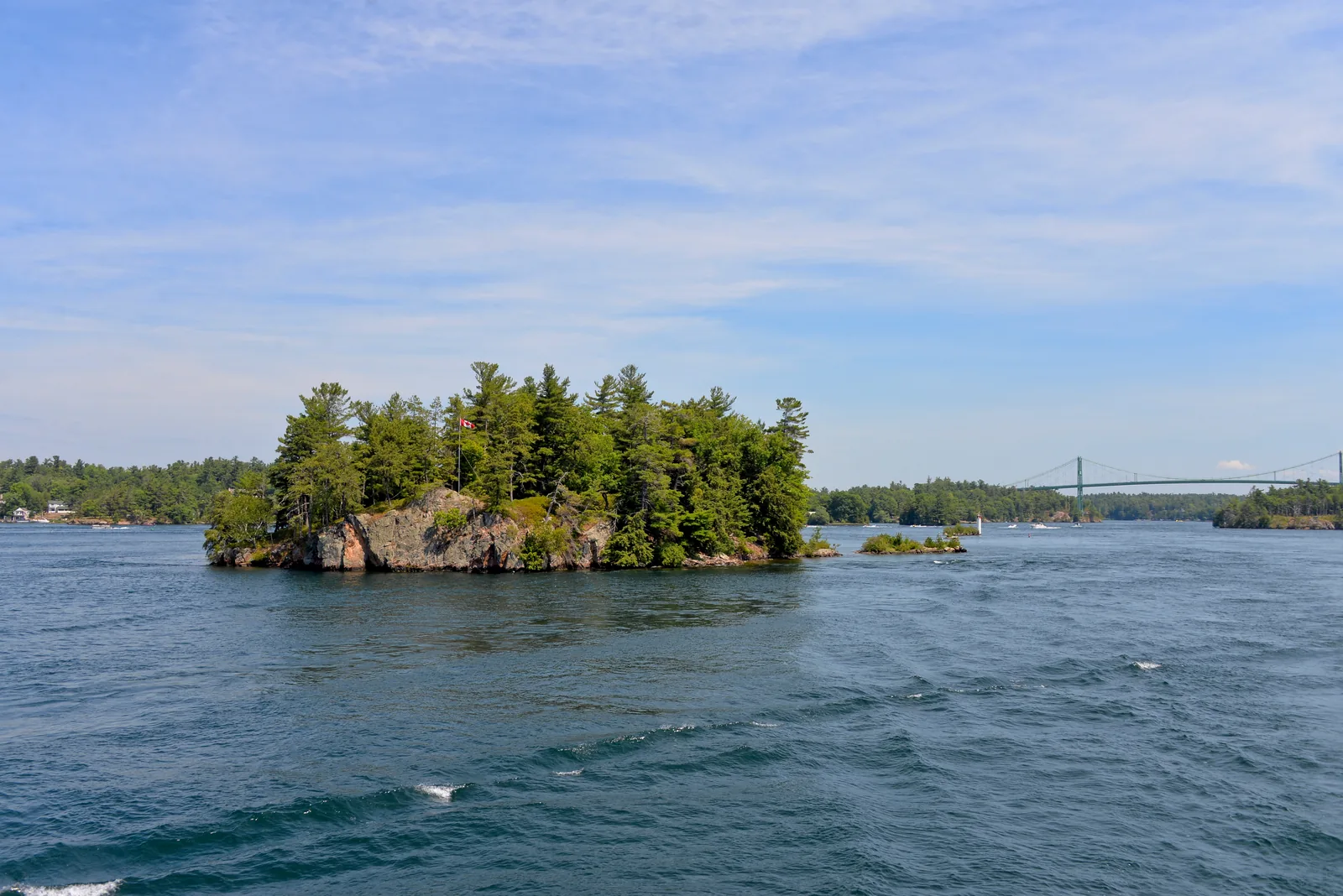 A small forested granite island with a Canadian flag in the river, a suspension bridge in the background on the right