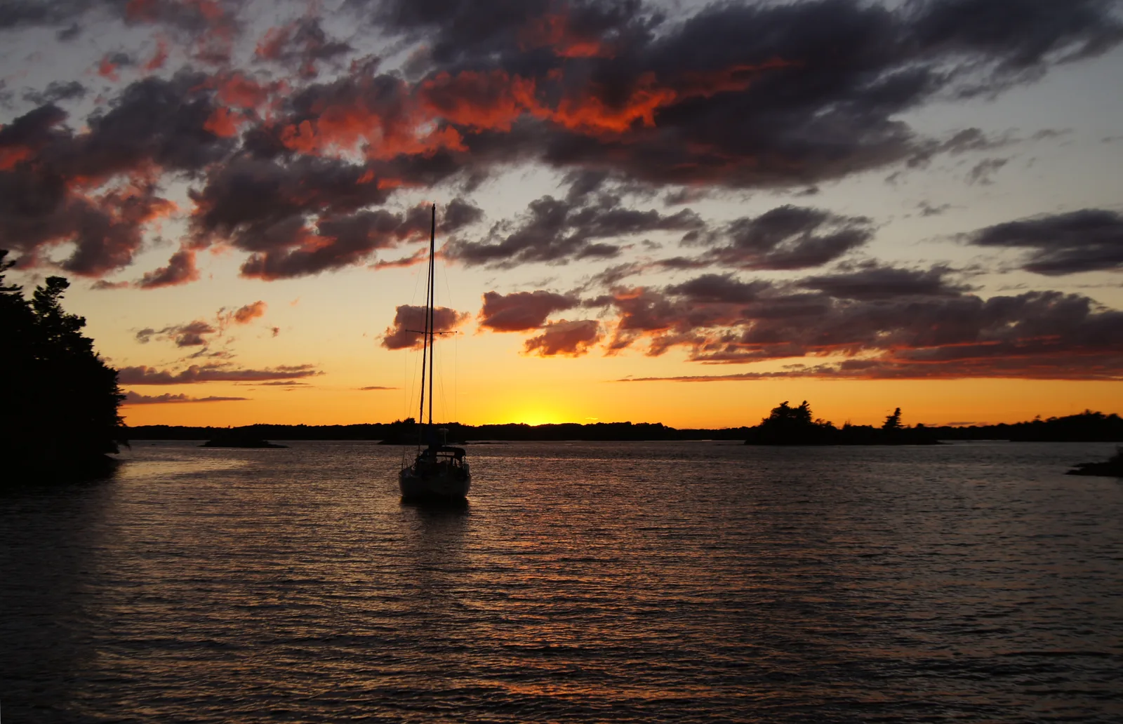 A sailboat lies off a forested island at sunset, the sky clouded orange and violet