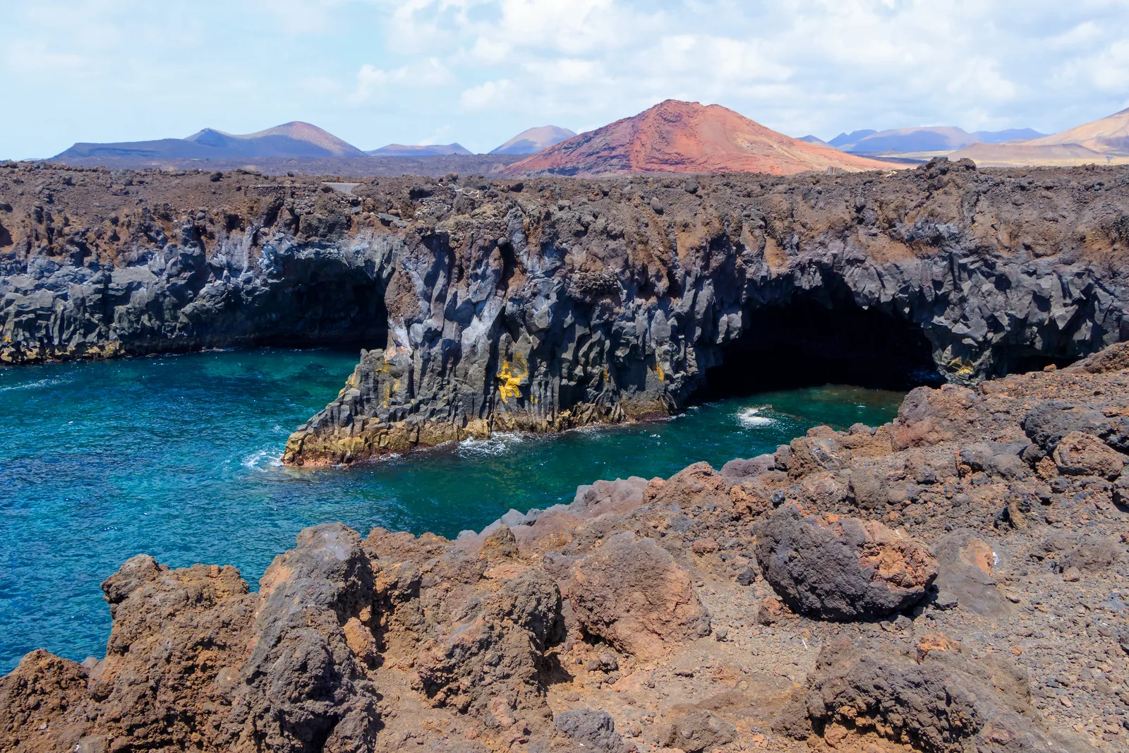 Turquoise surf in the black lava cliffs of Los Hervideros, with red volcanic cones behind