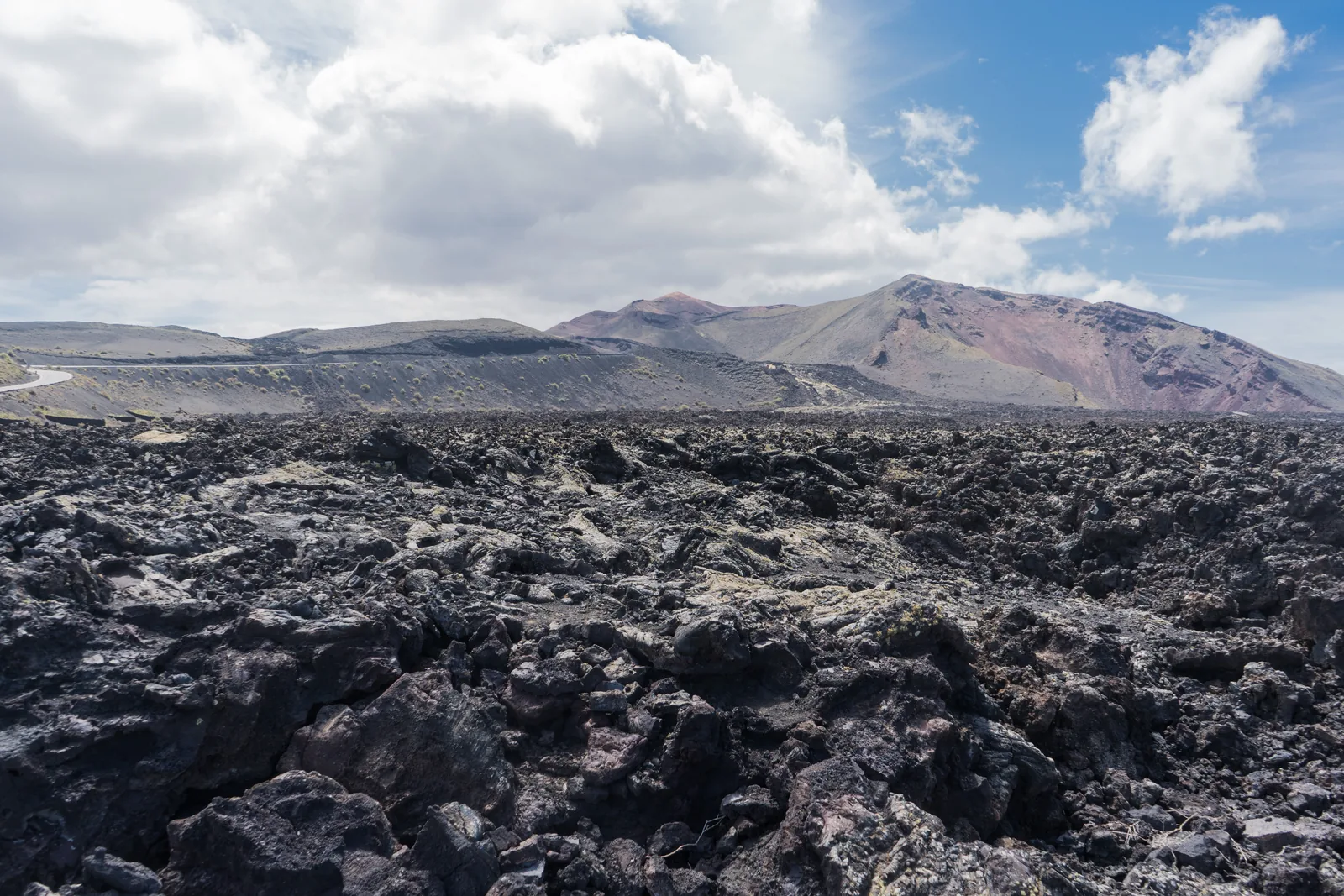 A black aa lava field stretching to the horizon, with the colourful volcanic cones of Timanfaya National Park under cloud behind
