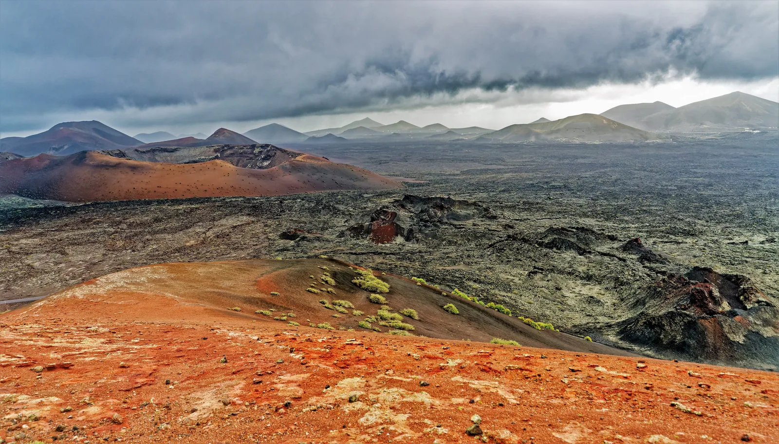 Red and black volcanic cones of Timanfaya National Park under dark cloud, with lapilli and the first lichens in the foreground