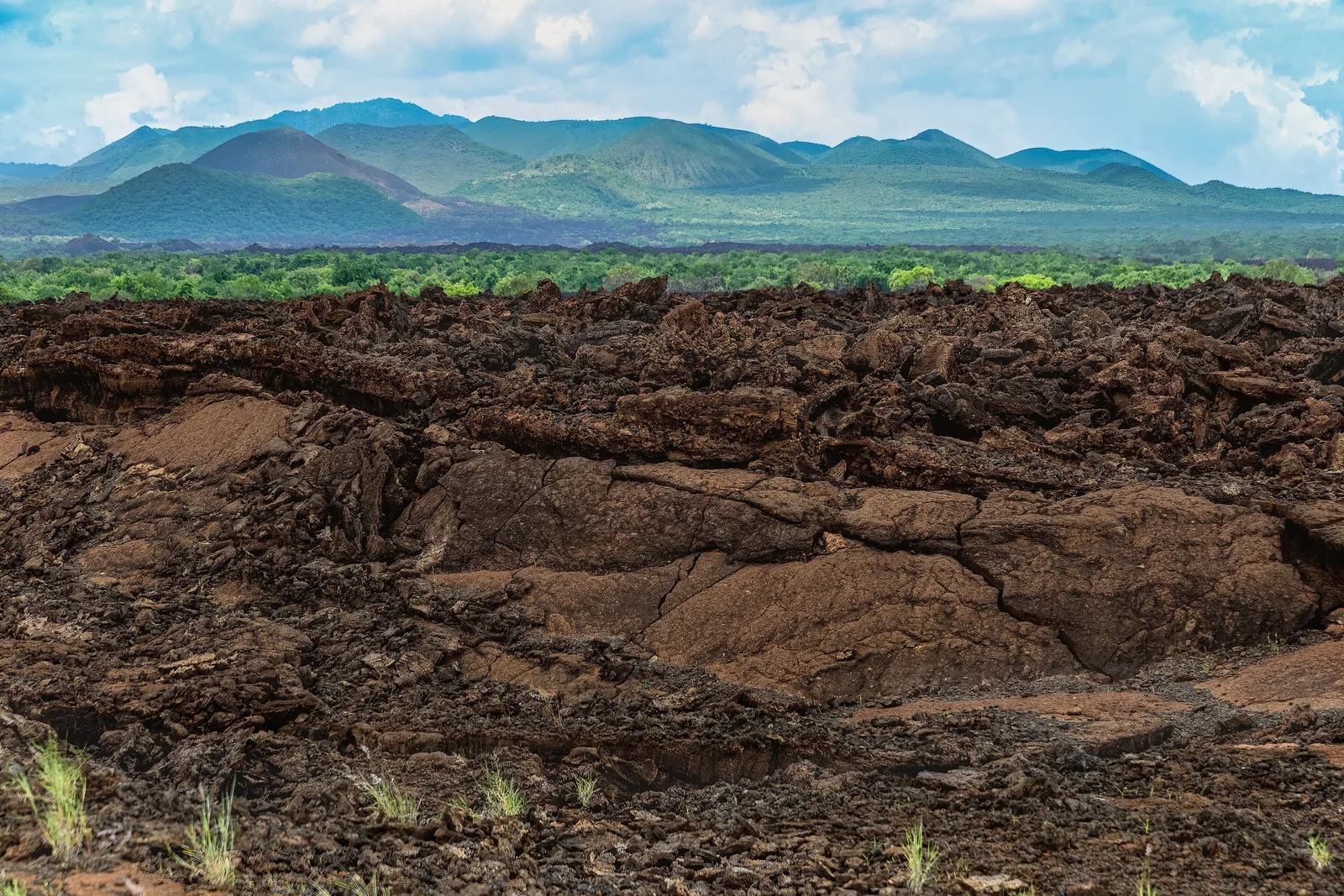 The black, jagged lava field of Shetani in Tsavo West National Park, with green hills under a cloudy sky behind