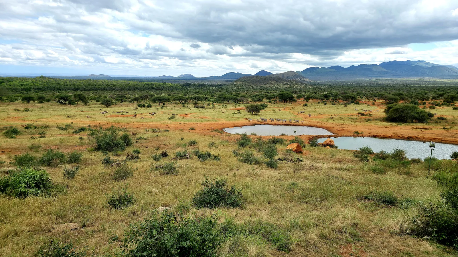 Wide bushland in Tsavo West National Park with two waterholes, hills and mountains under clouds behind