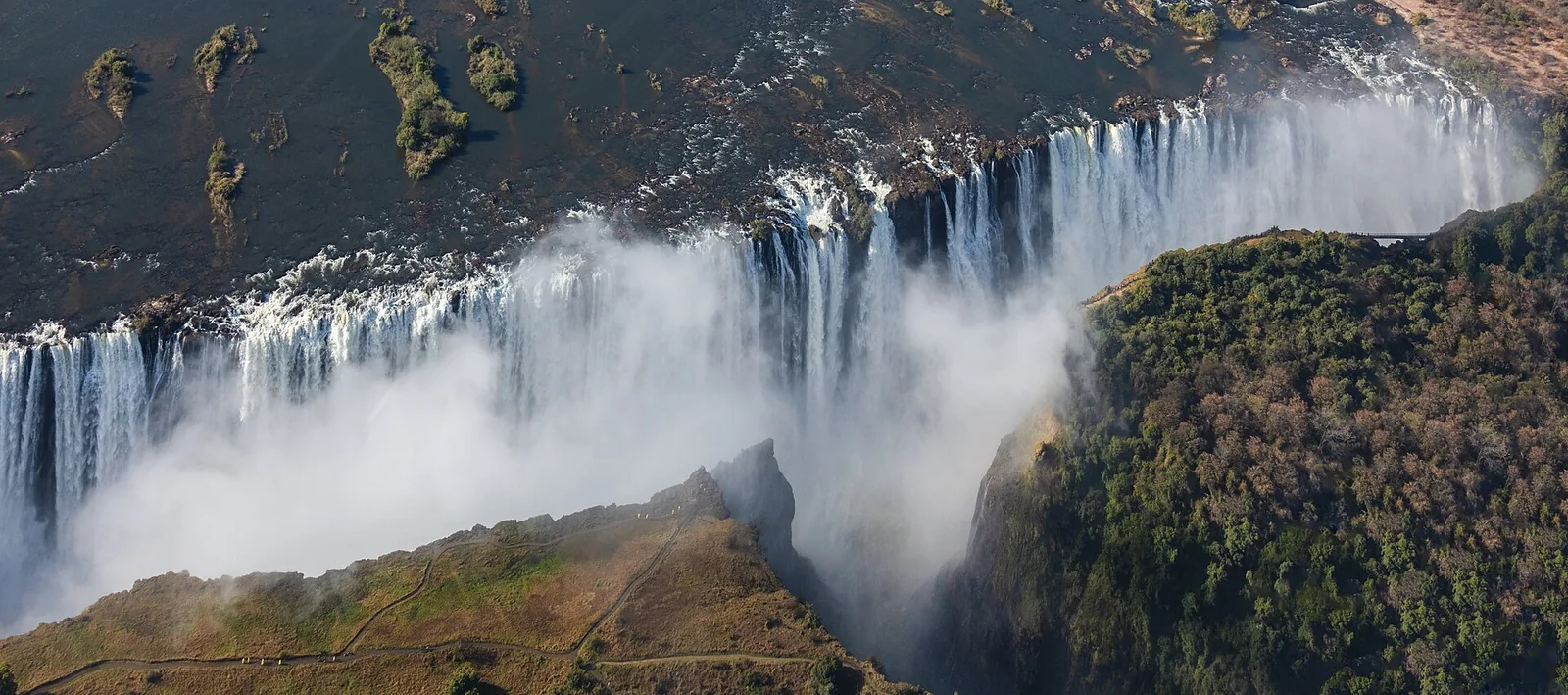 Aerial view of Victoria Falls: the Zambezi plunges along a broad front into the first gorge, spray rising, with the green rainforest on the Zimbabwean side to the right