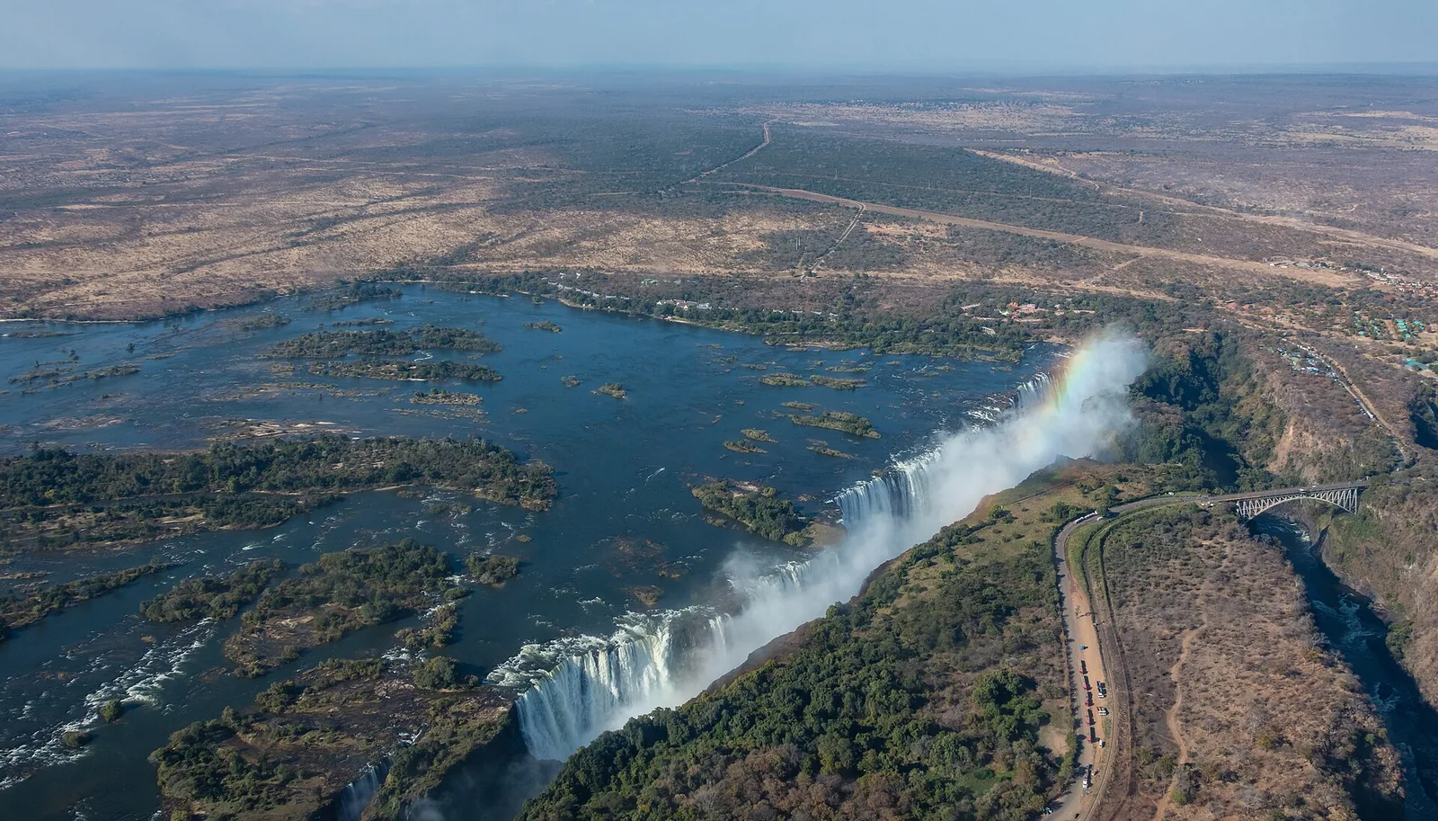 Aerial view of Victoria Falls with the Zambezi and its islands above the edge, water plunging into the gorge and the road along the bank