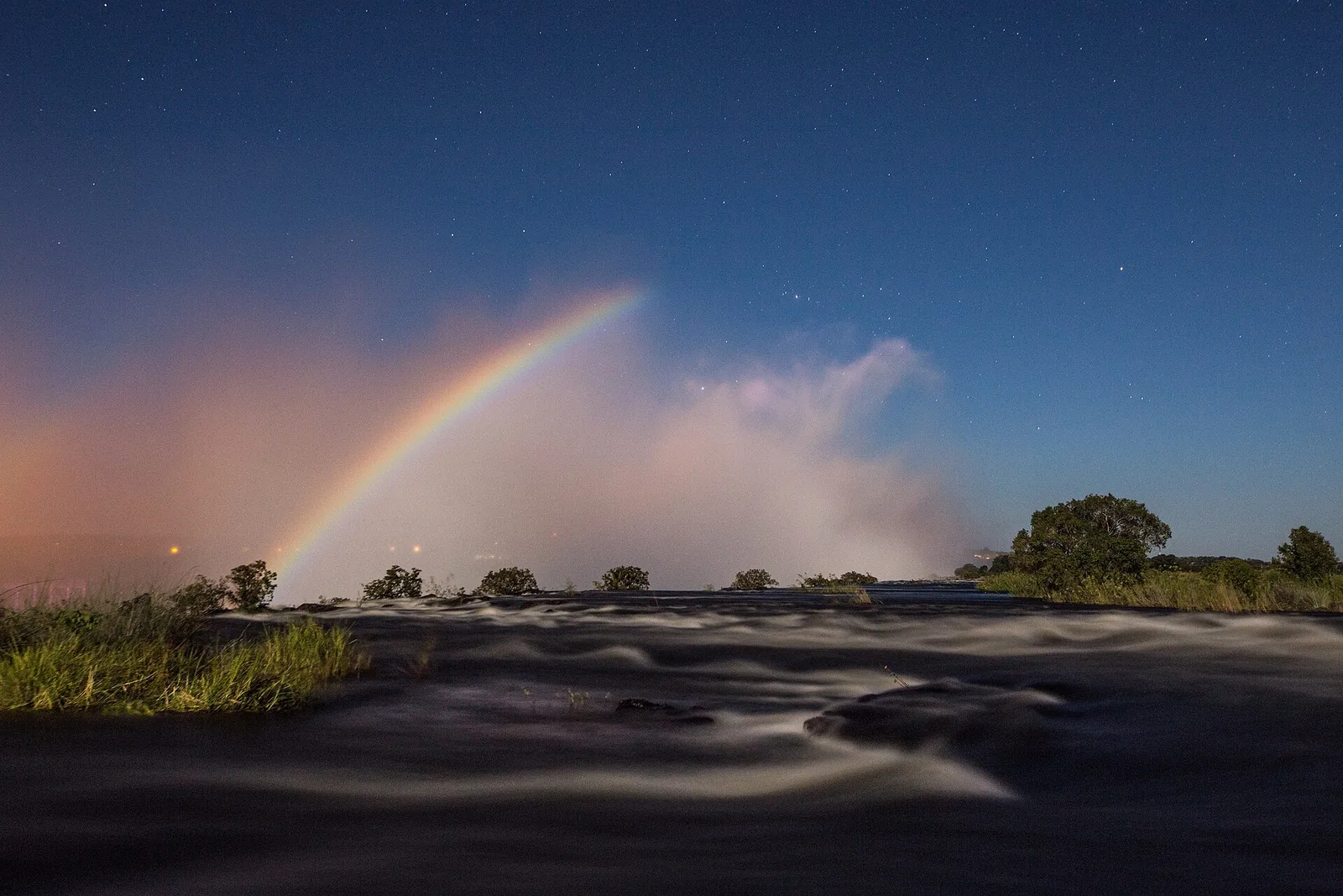 A lunar rainbow arches over the spray of Victoria Falls at night, below it the dark Zambezi and stars in the sky