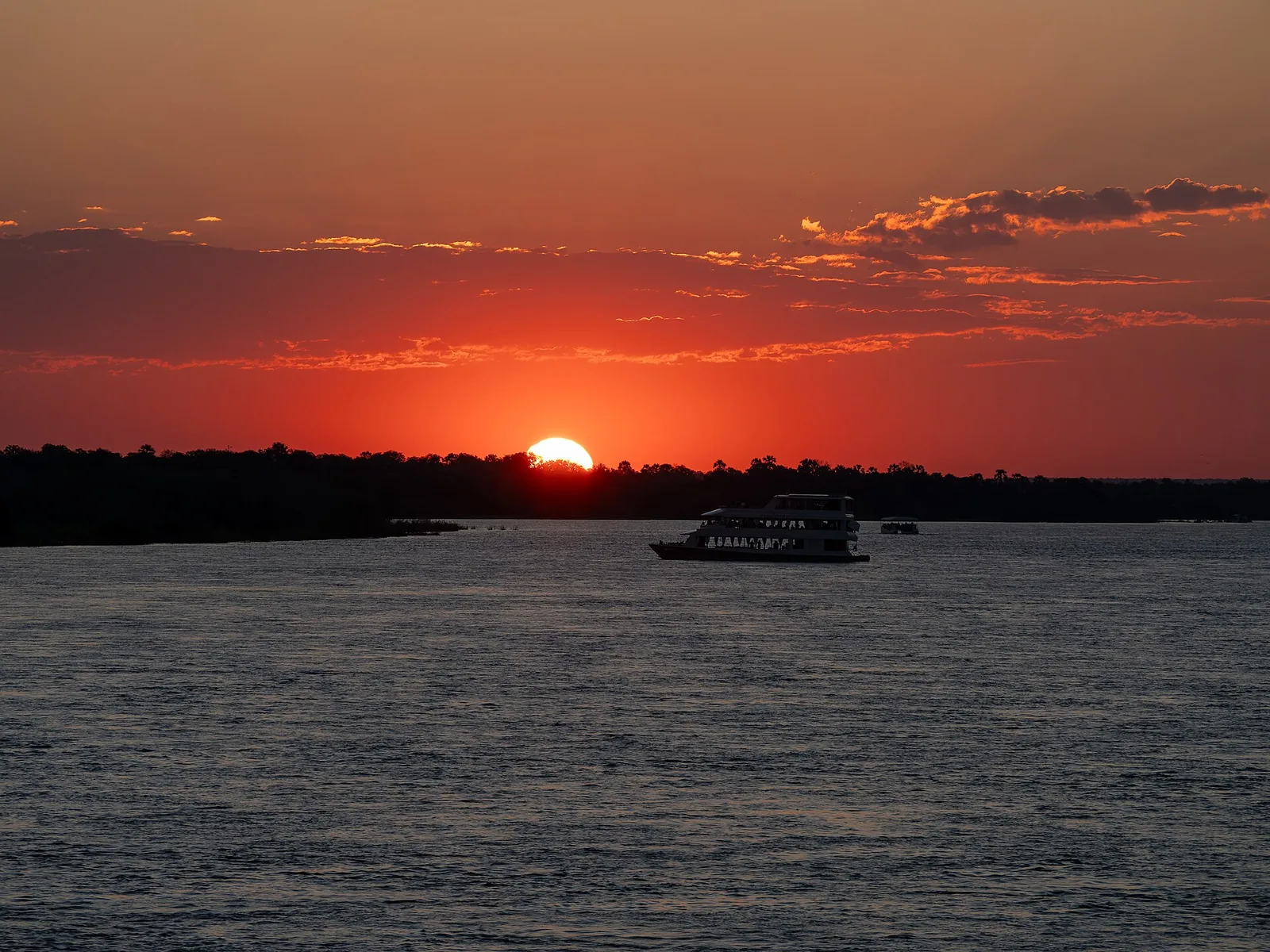 Sunset on the Zambezi above Victoria Falls, a tour boat in front of the dark bank under a red sky