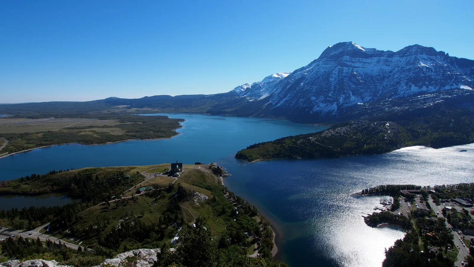 View from Bear's Hump in Waterton Lakes National Park across Upper Waterton Lake: flat prairie on the left, the snow-streaked mountain wall on the right, the Prince of Wales Hotel on the rise