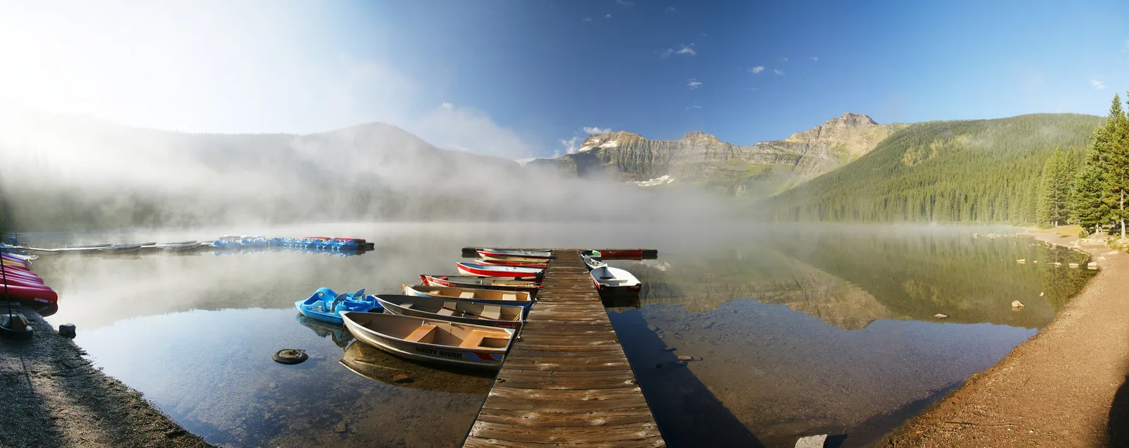 Early morning at Cameron Lake in Waterton Lakes National Park: mist over the water, a dock with a row of canoes, forested mountain slopes behind