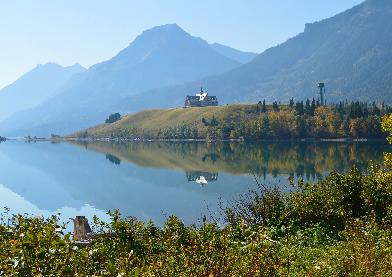 The Prince of Wales Hotel in Waterton Lakes National Park on a rise above the mirror-smooth Waterton lake, mountains behind, autumn shrubs on the shore in front