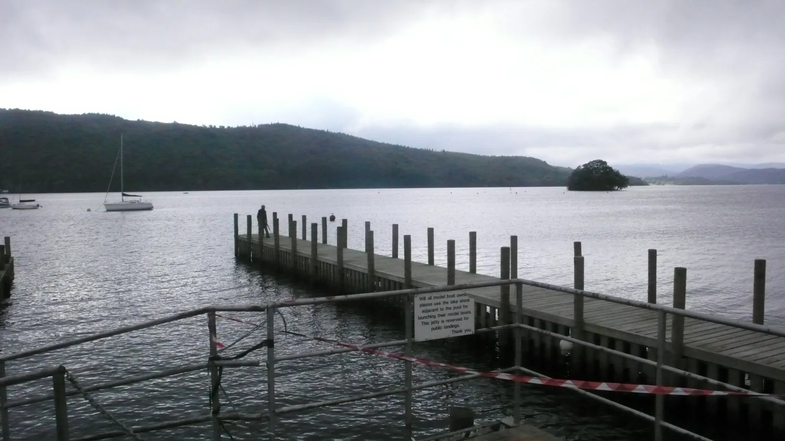 A wooden jetty reaching into Windermere in the Lake District, with a small island and hills under cloud behind