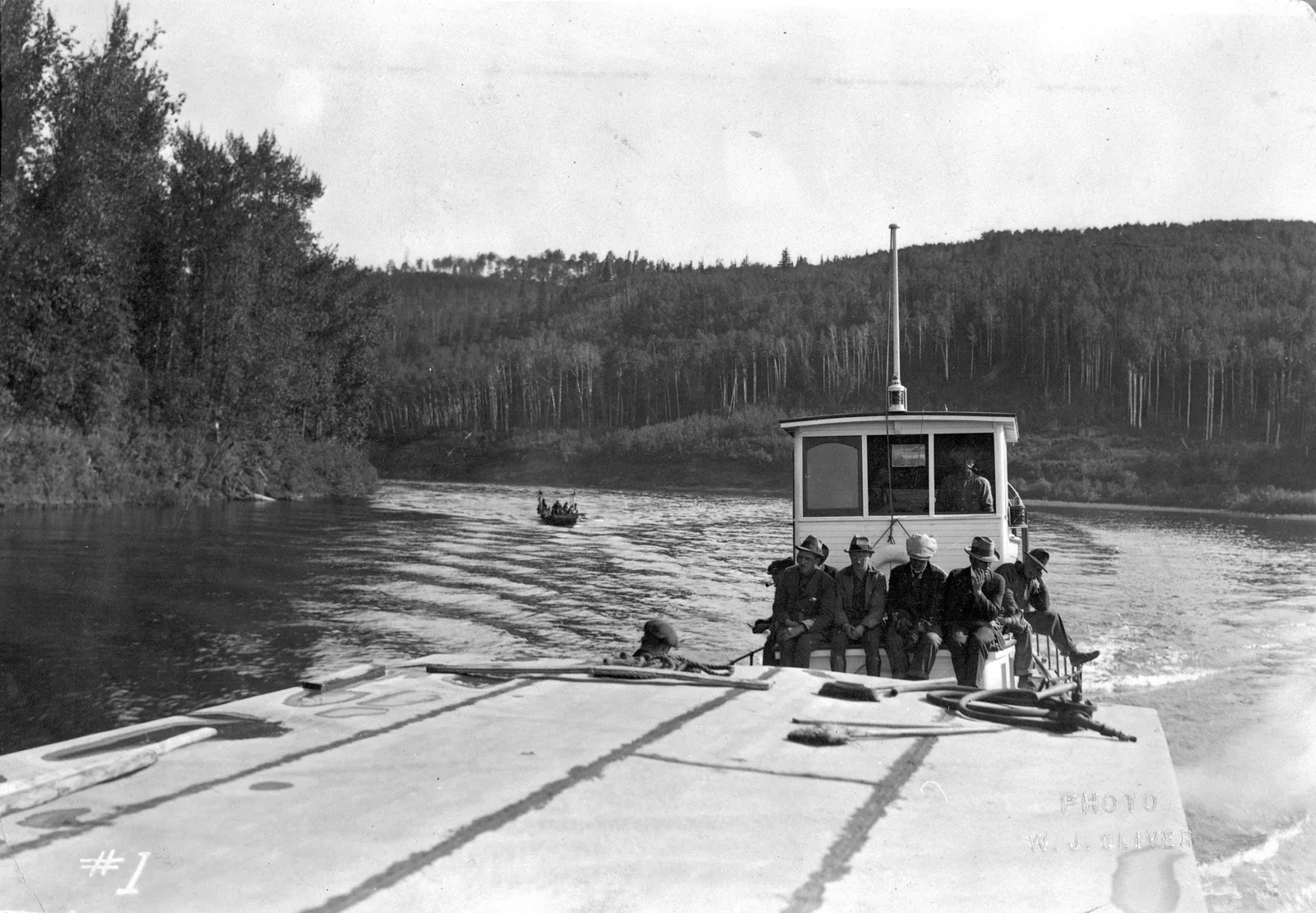 Historic black-and-white photo from Wood Buffalo National Park: a tugboat with men on deck pushes a barge across a river between forested banks