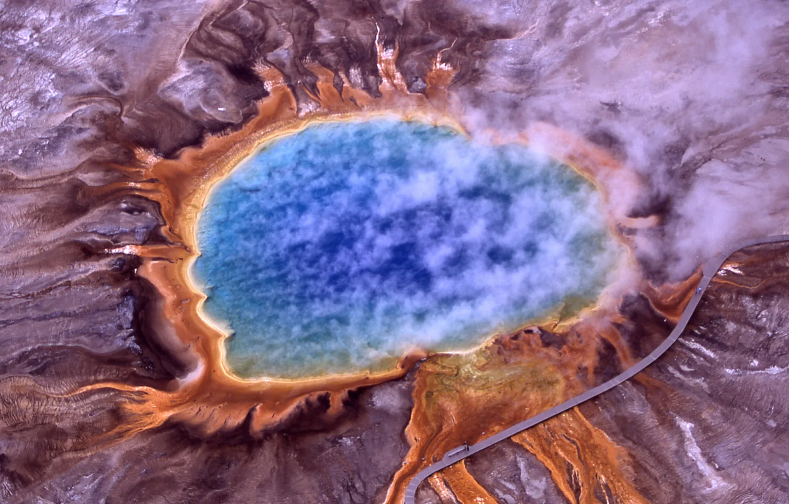 Aerial view of a circular hot spring in Yellowstone National Park with a blue centre and orange-yellow edges amid pale sinter flats.