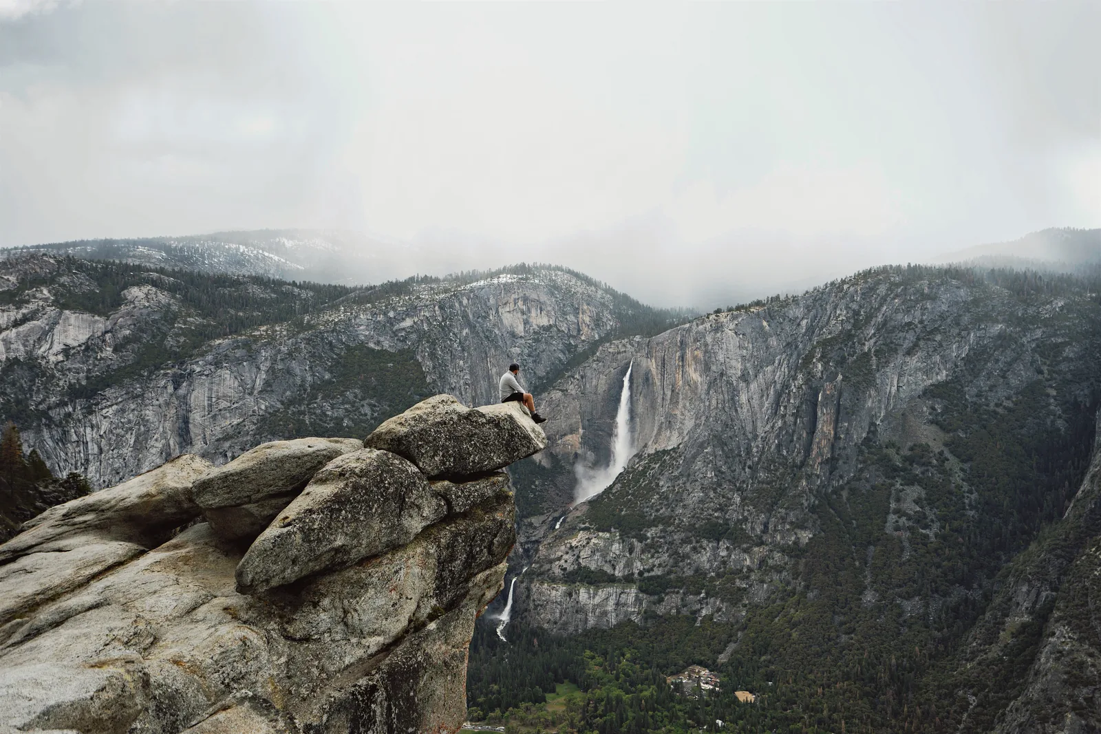 A person sits on a jutting rock at Glacier Point in Yosemite National Park; opposite, Yosemite Fall plunges down the valley wall