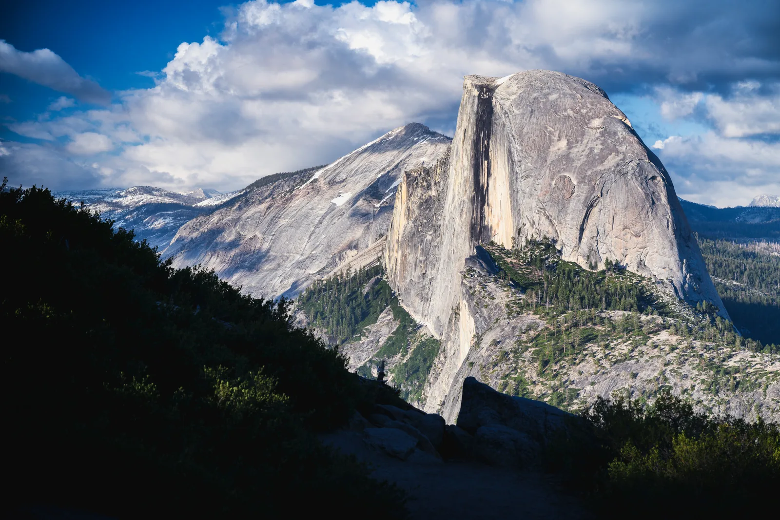 Half Dome in Yosemite National Park in evening light: a sheer, split granite dome above wooded slopes, with snow-patched mountains behind
