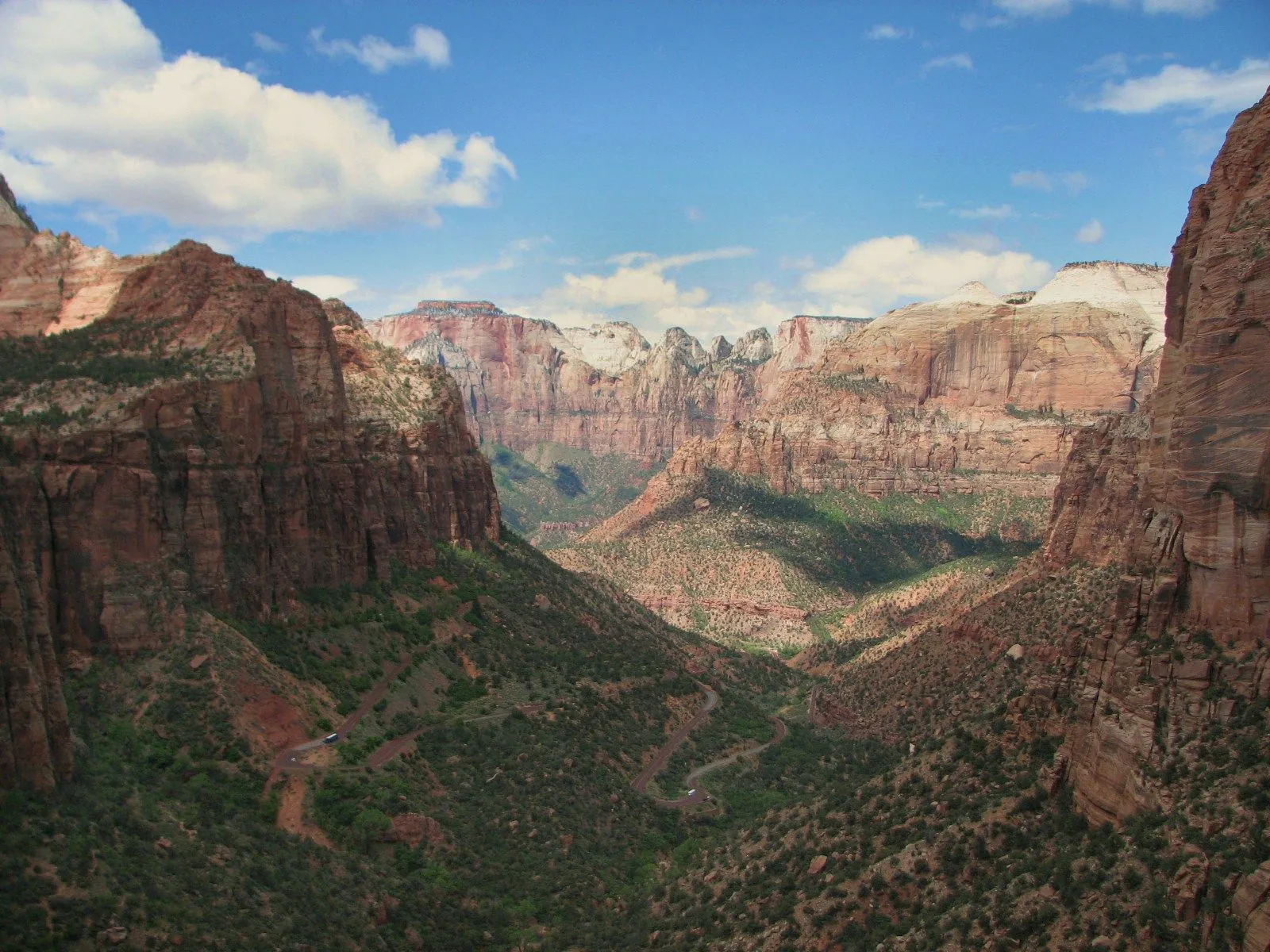 A deep canyon in Zion National Park between red sandstone walls, the switchbacks of a road below and pale peaks behind under cumulus clouds.