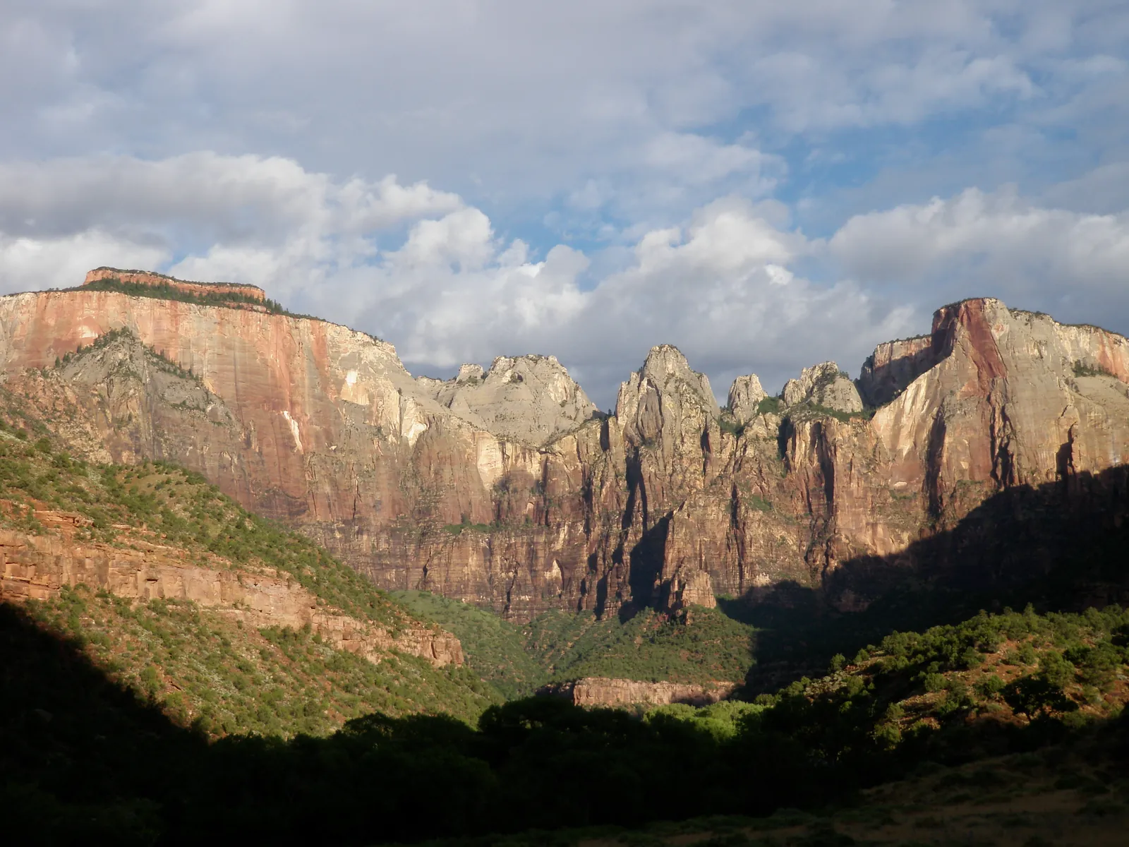 The Towers of the Virgin in Zion National Park in the first sunlight: red and pale sandstone walls above a dark wooded canyon floor, with clouds above.